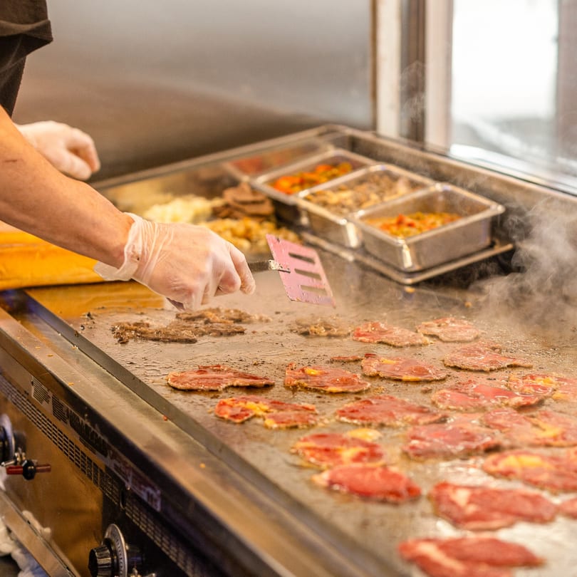 A gloved hand uses a spatula to cook thin slices of steak on a steaming hot griddle.