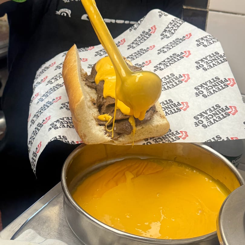 A Steve's Prince of Steaks cheesesteak being drenched in bright yellow cheese sauce from a ladle.